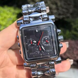 May include: A silver-toned Oakley watch with a square black face. The watch features red and white hands, multiple dials, and a date display. The watchband is made of the same silver material as the watch case.