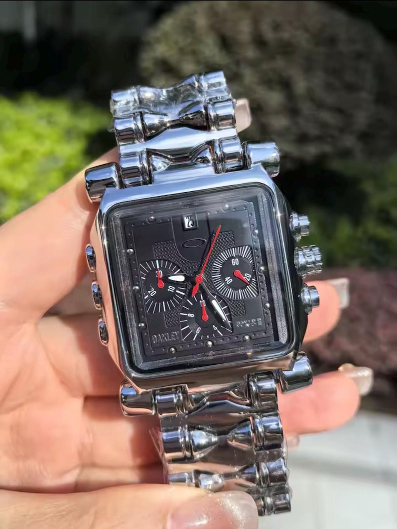 May include: A silver-toned Oakley watch with a square black face. The watch features red and white hands, multiple dials, and a date display. The watchband is made of the same silver material as the watch case.