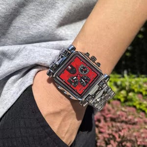 May include: A silver-toned wristwatch with a square red face and multiple sub-dials. The watch has a detailed, industrial design with a stainless steel band. The watch is on a wrist.