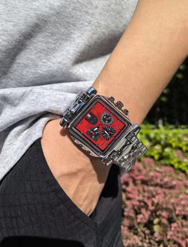 May include: A silver-toned wristwatch with a square red face and multiple sub-dials. The watch has a detailed, industrial design with a stainless steel band. The watch is on a wrist.