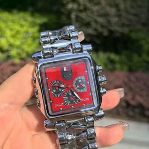 May include: A silver-toned wristwatch with a square red face and black sub-dials. The watch features a silver link band and is held against a blurred green background. The watch face has the brand name "Oakley".
