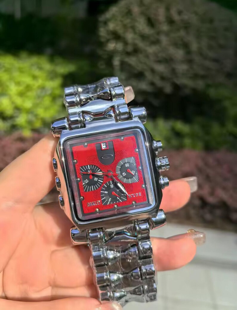 May include: A silver-toned wristwatch with a square red face and black sub-dials. The watch features a silver link band and is held against a blurred green background. The watch face has the brand name "Oakley".