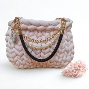 Könnte beinhalten: Eine hellrosa, handgestrickte Handtasche mit einem schwarzen Ledergriff und goldfarbener Hardware. Die Tasche hat eine dekorative Kette und einen Perlenriemen sowie einen kleinen Anhänger. Eine rosa Blume liegt neben der Tasche.