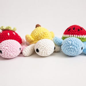 Pode incluir: Três tartarugas de pelúcia de crochê feitas à mão. Uma é rosa com uma casca de morango vermelha, uma é branca com uma casca de banana amarela e uma é azul com uma casca de melancia. Cada uma tem olhos pretos.