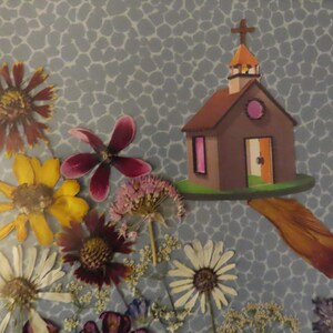 Pu&ograve; includere: Un collage incorniciato con una chiesa di carta con una croce, circondata da fiori pressati di vari colori. Lo sfondo &egrave; una carta blu a motivi e uno spartito musicale &egrave; visibile.