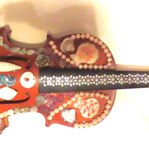 May include: A decorative violin with a reddish-brown body, adorned with various seashells, pearls, and a starfish. The neck is black, embellished with white accents. The violin's design incorporates a turtle and other sea-themed elements.