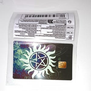 May include: A rectangular black card featuring a central white sun-like symbol with an embedded pentagram. The card has a gold-colored chip on the right side and a background with faint blue and purple geometric patterns and occult-style symbols.