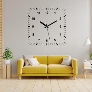 May include: A black and white clock decal with Roman numerals on a white wall. The clock is above a yellow sofa with two pillows.