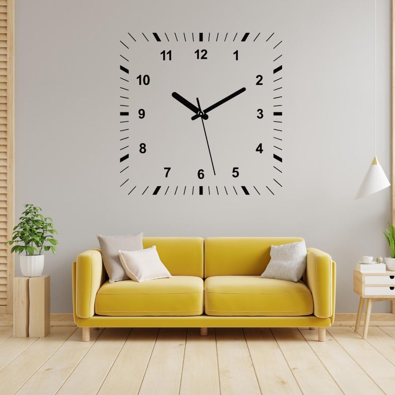 May include: A black and white clock decal with Roman numerals on a white wall. The clock is above a yellow sofa with two pillows.