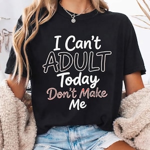 May include: A black short-sleeved t-shirt featuring the white text "I Can't ADULT Today" and the pinkish-white text "Don't Make Me" printed on the front. The shirt has a crew neckline and is worn with blue denim shorts and a beige textured cardigan.