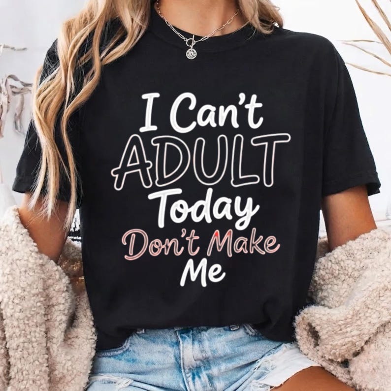May include: A black short-sleeved t-shirt featuring the white text "I Can't ADULT Today" and the pinkish-white text "Don't Make Me" printed on the front. The shirt has a crew neckline and is worn with blue denim shorts and a beige textured cardigan.