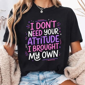 May include: A black short-sleeved t-shirt featuring the white, pink, and purple text "I DON'T NEED YOUR ATTITUDE I BROUGHT MY OWN" printed on the front. The graphic design includes small star and dash accents surrounding the bold, stylized lettering.