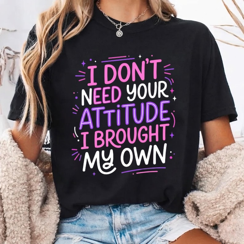 May include: A black short-sleeved t-shirt featuring the white, pink, and purple text "I DON'T NEED YOUR ATTITUDE I BROUGHT MY OWN" printed on the front. The graphic design includes small star and dash accents surrounding the bold, stylized lettering.