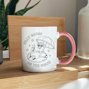 May include: White ceramic mug with a pink handle and interior. The mug features a black and white illustration of a goose wearing sunglasses and holding an ice cream cone under an umbrella. Text on the mug reads "Silly Goose on the Beach."