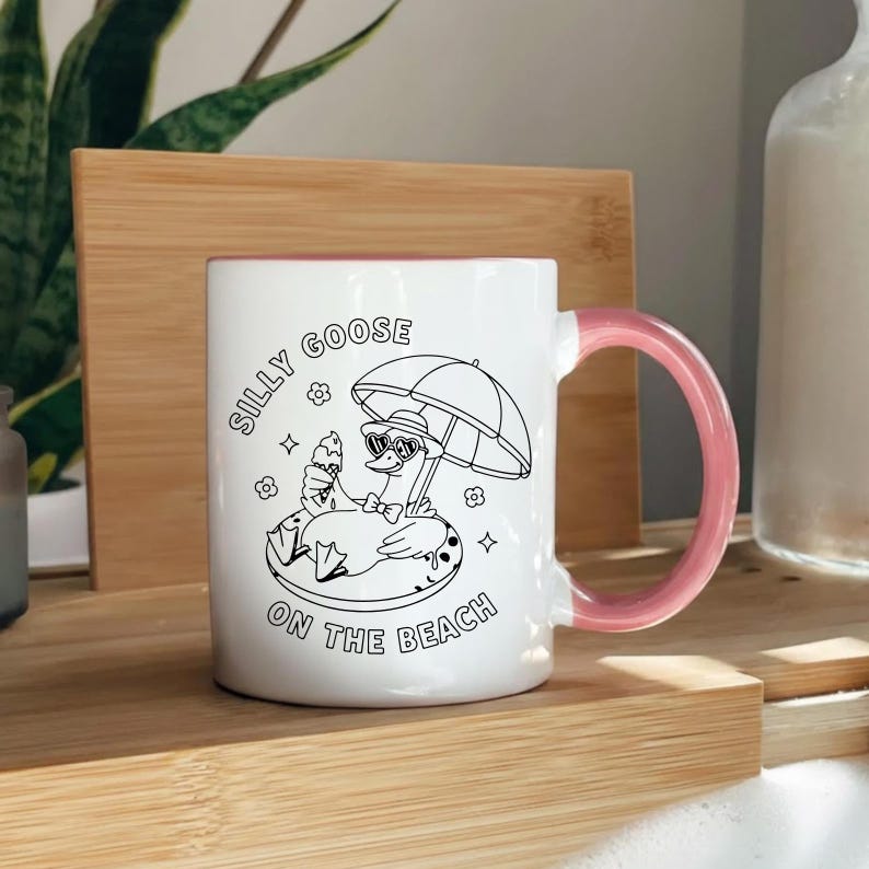 May include: White ceramic mug with a pink handle and interior. The mug features a black and white illustration of a goose wearing sunglasses and holding an ice cream cone under an umbrella. Text on the mug reads "Silly Goose on the Beach."