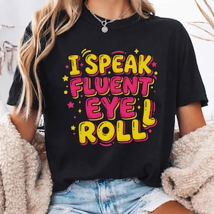 May include: A black short-sleeved t-shirt featuring the text I SPEAK FLUENT EYE ROLL in bold yellow and pink lettering. The design includes small yellow stars and dots scattered around the words, creating a vibrant graphic on the front of the shirt.