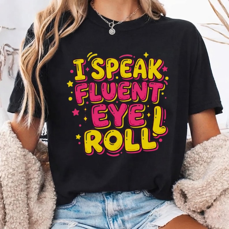 May include: A black short-sleeved t-shirt featuring the text I SPEAK FLUENT EYE ROLL in bold yellow and pink lettering. The design includes small yellow stars and dots scattered around the words, creating a vibrant graphic on the front of the shirt.