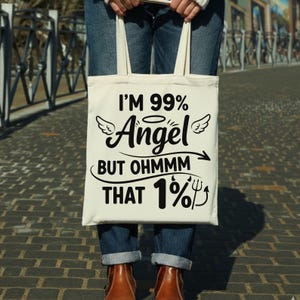 May include: Cream-colored tote bag with the text "I'M 99% Angel BUT OHMMM THAT 1%" in black. The bag has a humorous design with angel wings and a devil's pitchfork. The bag is held by a person wearing blue jeans and brown boots.