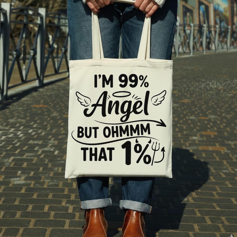 May include: Cream-colored tote bag with the text "I'M 99% Angel BUT OHMMM THAT 1%" in black. The bag has a humorous design with angel wings and a devil's pitchfork. The bag is held by a person wearing blue jeans and brown boots.