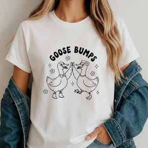 May include: White t-shirt with a black graphic that reads "GOOSE BUMPS" above two cartoon ducks giving each other a high five. The ducks are outlined in black, with one wearing a bow tie and boots, and the other wearing a flower crown and necklace.
