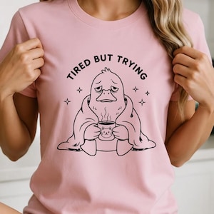 May include: Pink t-shirt with the text "TIRED BUT TRYING" above a black and white illustration of a duck-like creature wrapped in a blanket, holding a mug with a cat design. Four small star shapes surround the illustration.