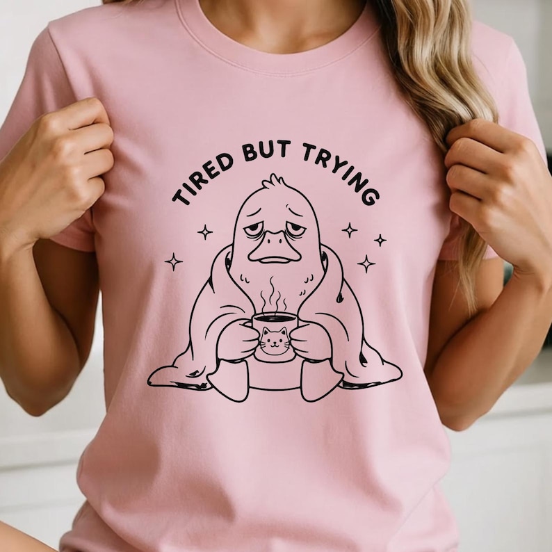 May include: Pink t-shirt with the text "TIRED BUT TRYING" above a black and white illustration of a duck-like creature wrapped in a blanket, holding a mug with a cat design. Four small star shapes surround the illustration.