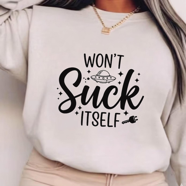 May include: A light beige sweatshirt with the text "WON'T Suck ITSELF" in black. The design includes a UFO, stars, and a key. The sweatshirt is a casual, comfortable style.