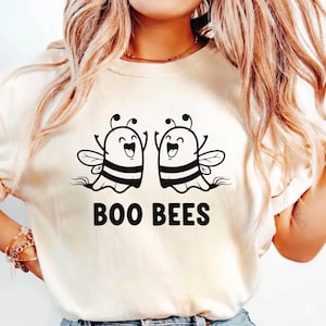 May include: Cream-colored t-shirt featuring a graphic of two cartoon bees with smiling faces and raised arms. The bees have black and yellow striped bodies and wings. Below the bees, the text "BOO BEES" is printed in black.