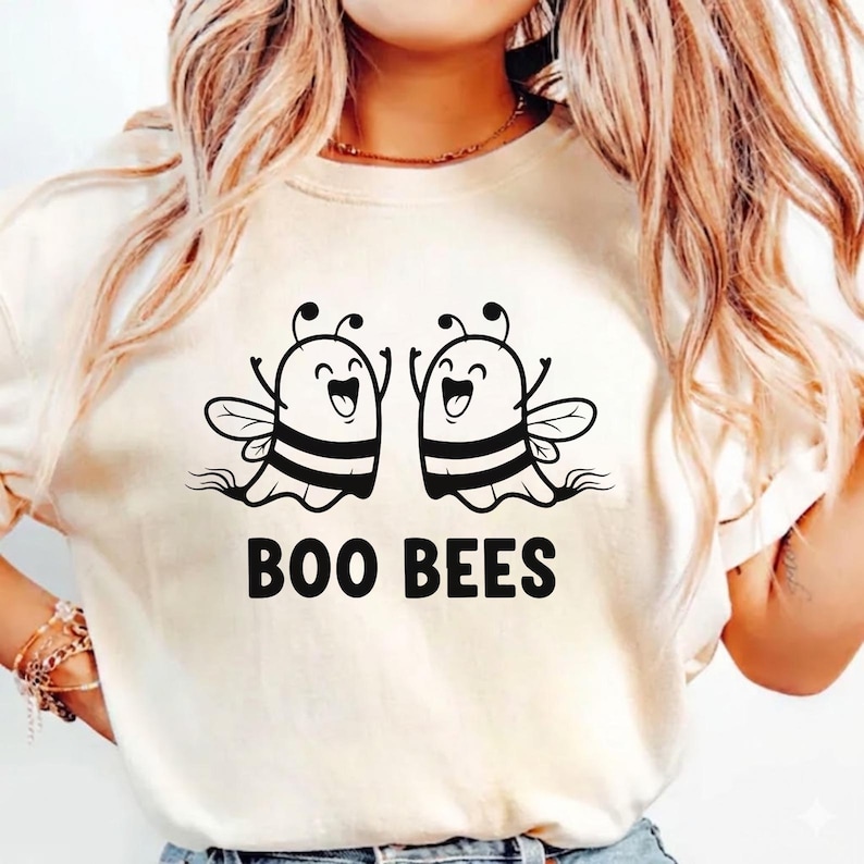 May include: Cream-colored t-shirt featuring a graphic of two cartoon bees with smiling faces and raised arms. The bees have black and yellow striped bodies and wings. Below the bees, the text "BOO BEES" is printed in black.