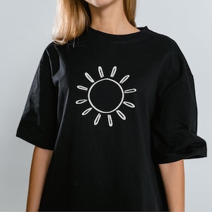 Może przedstawiać: Czarny t-shirt z białą grafiką słońca. Projekt słońca to prosty kontur z okrągłym środkiem i promienistymi liniami. Koszulka ma kr&oacute;tkie rękawy i okrągły dekolt.