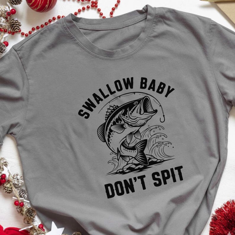 May include: A gray t-shirt featuring a black and white graphic of a fish jumping out of the water with a fishing hook. The text "SWALLOW BABY DON'T SPIT" is above and below the fish illustration.