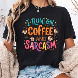 May include: A black t-shirt featuring a colorful graphic with the text I RUN ON COFFEE AND SARCASM. The design includes illustrations of four coffee mugs, scattered coffee beans, and small yellow stars, with the text in blue, orange, and pink.