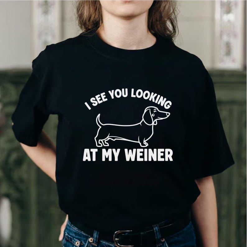 May include: Black t-shirt with white text and a dachshund illustration. The text reads "I SEE YOU LOOKING AT MY WEINER". The dachshund is drawn in a simple, cartoon style.