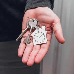 May include: A white house-shaped keychain with a clock face and black hands. The keychain is attached to a set of silver keys.
