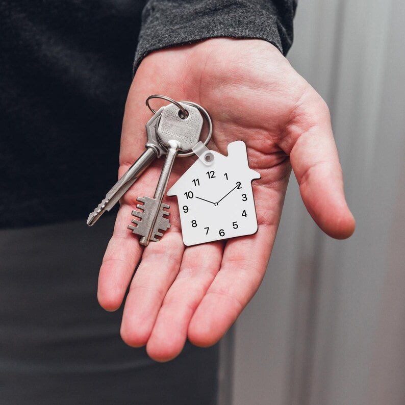 May include: A white house-shaped keychain with a clock face and black hands. The keychain is attached to a set of silver keys.