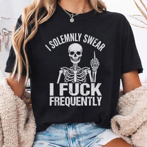May include: A black short-sleeve t-shirt featuring a white graphic of a skeleton giving the middle finger. The text above the skeleton reads I SOLEMNLY SWEAR and the text below reads I FUCK FREQUENTLY. The design has a distressed, vintage texture.