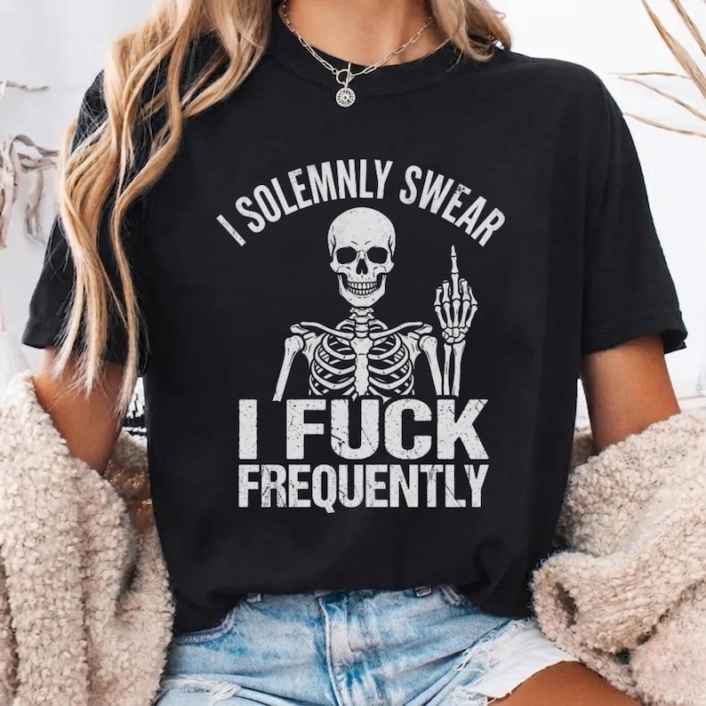May include: A black short-sleeve t-shirt featuring a white graphic of a skeleton giving the middle finger. The text above the skeleton reads I SOLEMNLY SWEAR and the text below reads I FUCK FREQUENTLY. The design has a distressed, vintage texture.