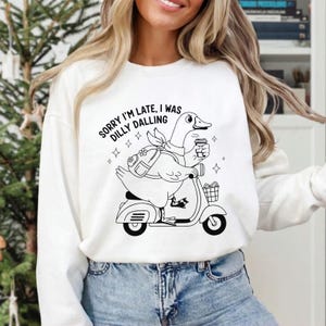 May include: White sweatshirt with a black and white graphic of a duck riding a scooter, holding a coffee cup, and wearing a backpack. The text "SORRY I'M LATE, I WAS DILLY DALLYING" is above the graphic.
