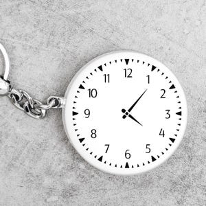 May include: A white round keychain with a clock face. The clock face has black numbers 1 through 12 and black hour and minute hands.