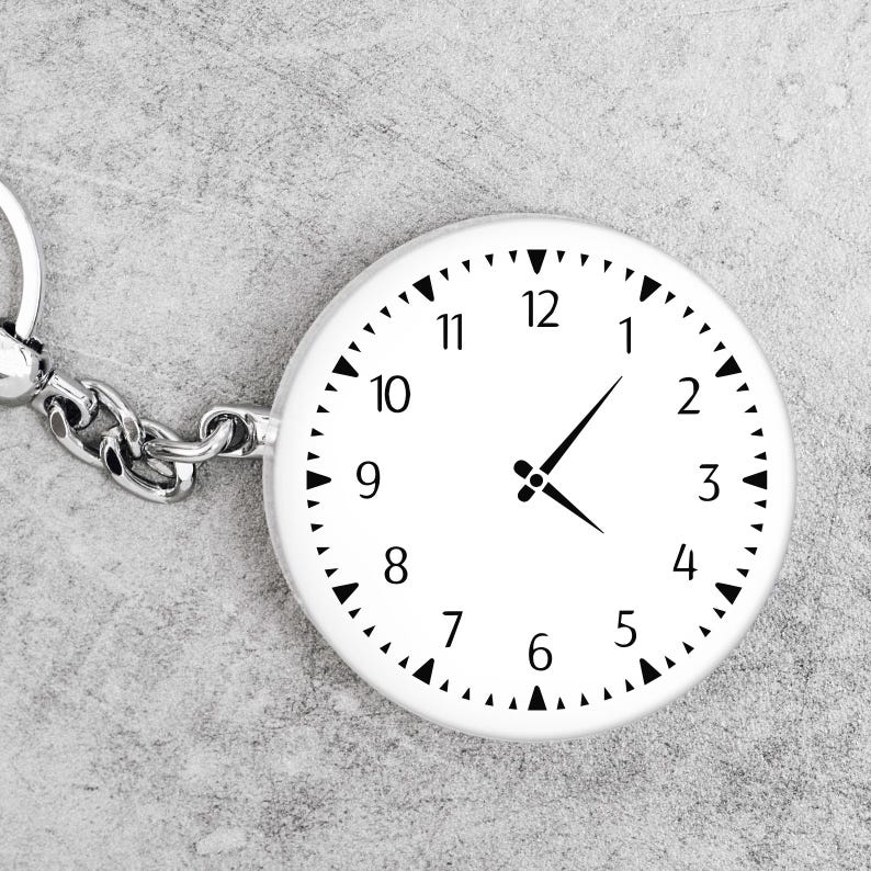 May include: A white round keychain with a clock face. The clock face has black numbers 1 through 12 and black hour and minute hands.