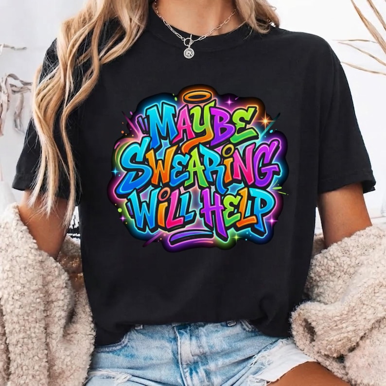 May include: A black short-sleeve t-shirt featuring a colorful graffiti-style graphic on the chest. The design displays the text "MAYBE SWEARING WILL HELP" in vibrant blue, purple, green, and orange letters with a small halo above the word "MAYBE".