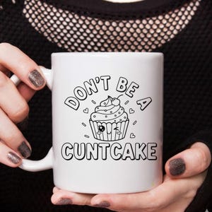 May include: White ceramic mug with the text "DON'T BE A CUNTCAKE" in black, along with a cupcake illustration. The mug has a white handle and is held by a person.