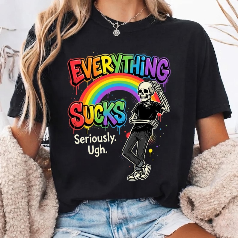 May include: A black t-shirt featuring a graphic of a skeleton standing under a rainbow. The text reads "EVERYTHING SUCKS Seriously. Ugh." in colorful, dripping letters. The skeleton wears a black shirt and pants with white sneakers.