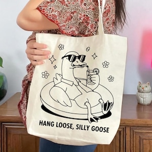 May include: Cream-colored tote bag featuring a black illustration of a goose wearing sunglasses and floating on an inner tube while drinking a beverage. The text "HANG LOOSE, SILLY GOOSE" is printed below the illustration. Floral and star graphics surround the goose.