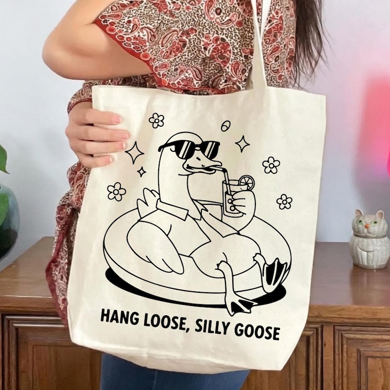 May include: Cream-colored tote bag featuring a black illustration of a goose wearing sunglasses and floating on an inner tube while drinking a beverage. The text "HANG LOOSE, SILLY GOOSE" is printed below the illustration. Floral and star graphics surround the goose.