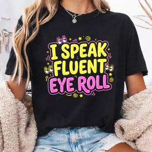 May include: A black t-shirt featuring the yellow text I SPEAK FLUENT and pink text EYE ROLL. The design includes cartoon eyes rolling in various directions, small yellow stars, and swirling accents, all set against a solid black fabric background.