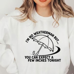 May include: White sweatshirt with a black graphic of an umbrella and the text "I'M NO WEATHERMAN BUT... YOU CAN EXPECT A FEW INCHES TONIGHT". The design is centered on the front of the shirt.
