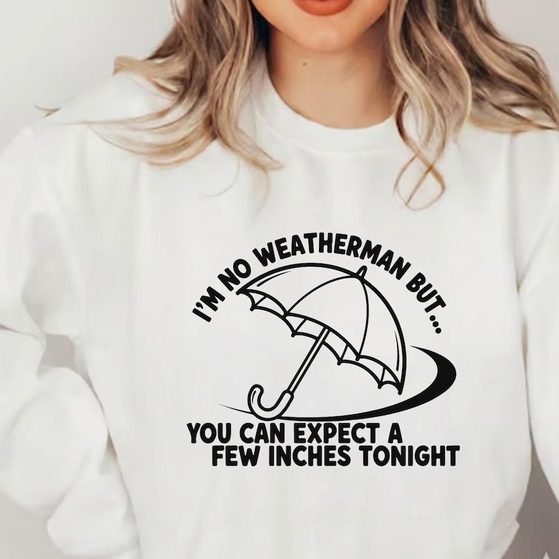 May include: White sweatshirt with a black graphic of an umbrella and the text "I'M NO WEATHERMAN BUT... YOU CAN EXPECT A FEW INCHES TONIGHT". The design is centered on the front of the shirt.