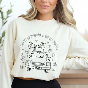 May include: Cream-colored sweatshirt with a black and white graphic. The graphic features a goose driving a car and holding a megaphone, surrounded by flowers and stars. Text reads "Honk if you're a silly goose."