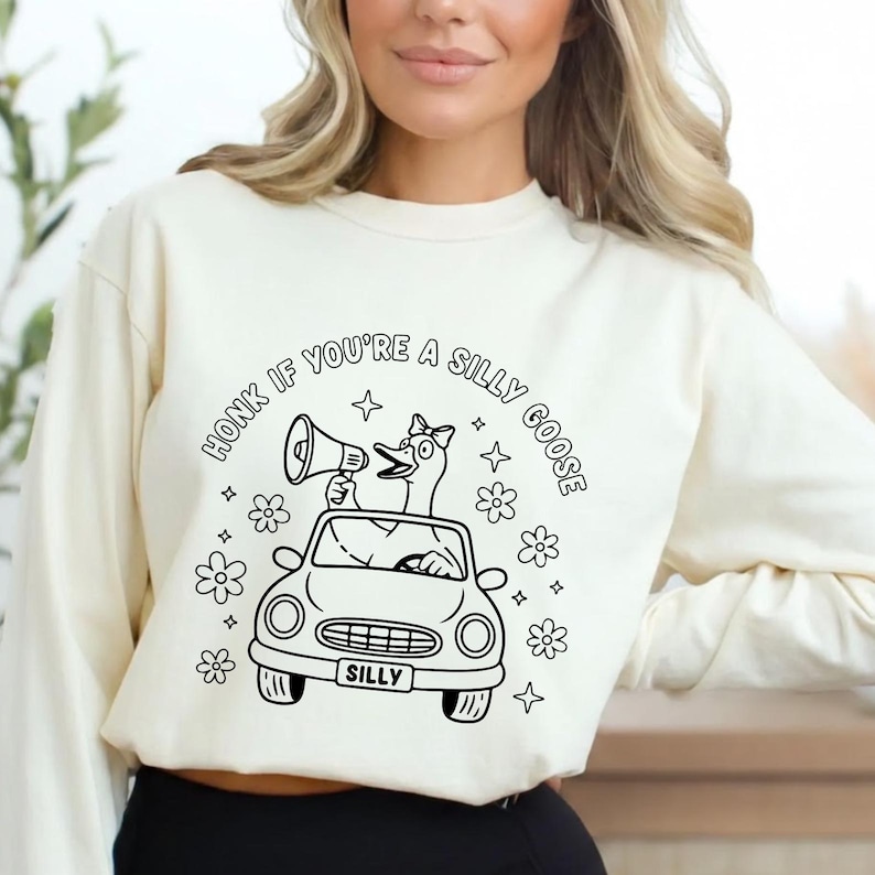 May include: Cream-colored sweatshirt with a black and white graphic. The graphic features a goose driving a car and holding a megaphone, surrounded by flowers and stars. Text reads "Honk if you're a silly goose."